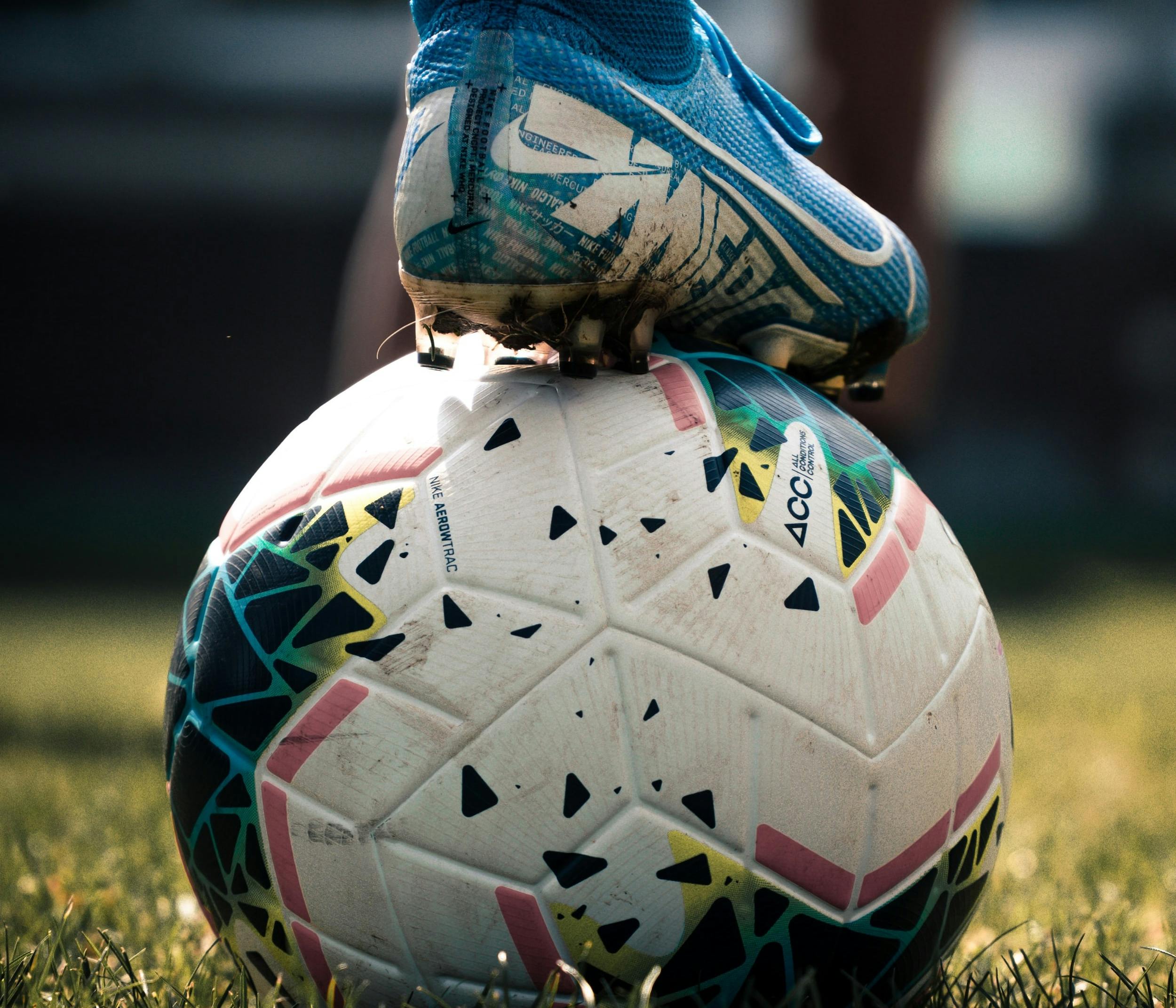 Piede di calciatore con scarpa blu a tacchetti appoggiato su un pallone da calcio su un campo erboso..