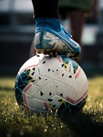 Piede di calciatore con scarpa blu a tacchetti appoggiato su un pallone da calcio su un campo erboso..