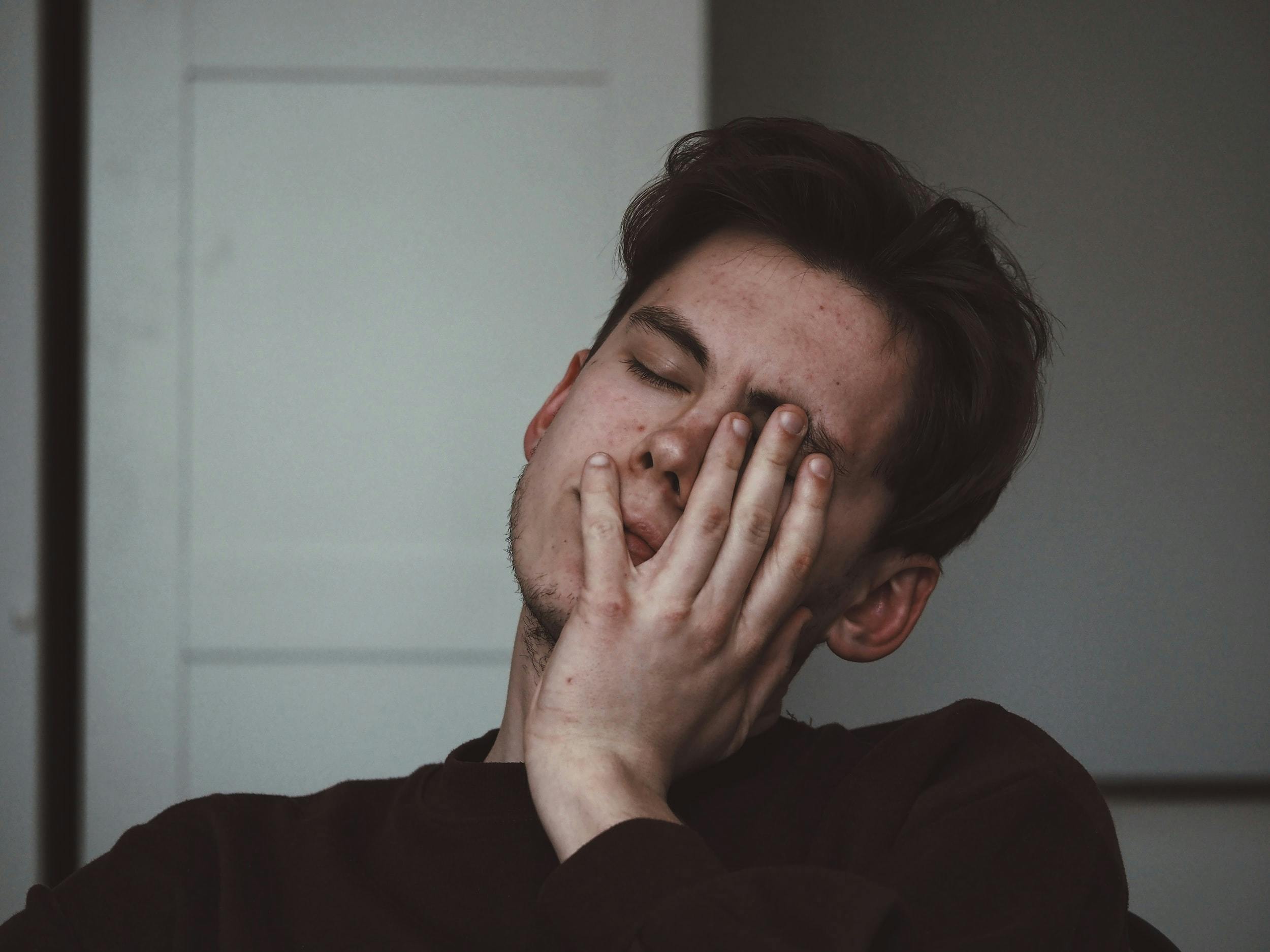 Foto di giovane ragazzo vestito con una maglia nera che si appoggia la mano sinistra sul volto chiudendo glio occhi, mostrando segni di stanchezza.