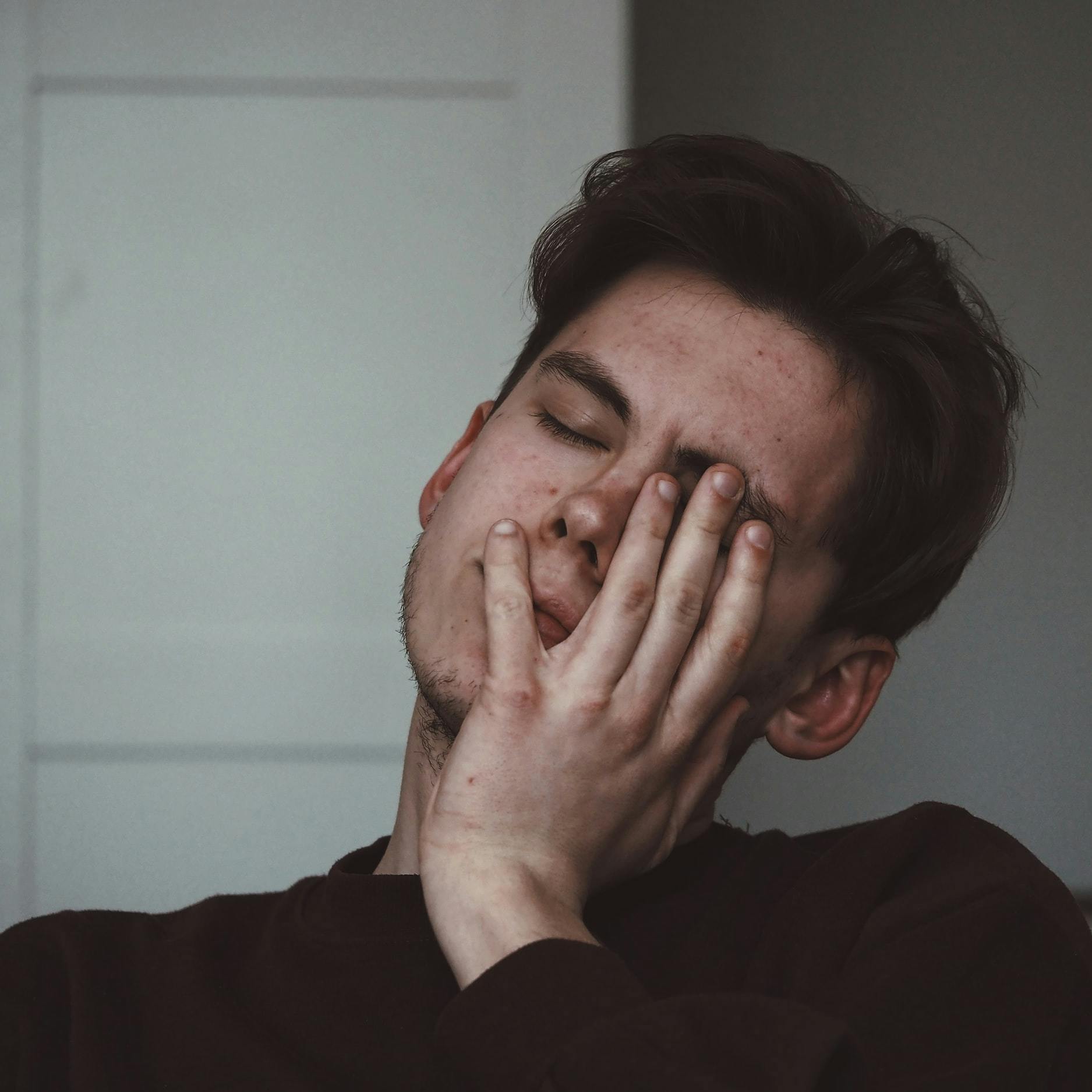 Foto di giovane ragazzo vestito con una maglia nera che si appoggia la mano sinistra sul volto chiudendo glio occhi, mostrando segni di stanchezza.