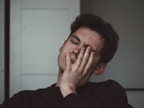 Young mandressed in a black shirt resting his left hand on his face closing his eyes, showing signs of fatigue.