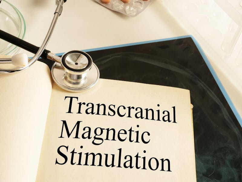 Pagina di libro con scritto "Transcranial Magnetic Stimulation" aperta su un tavolo su cui poggia uno stetoscopio e delle medicine.