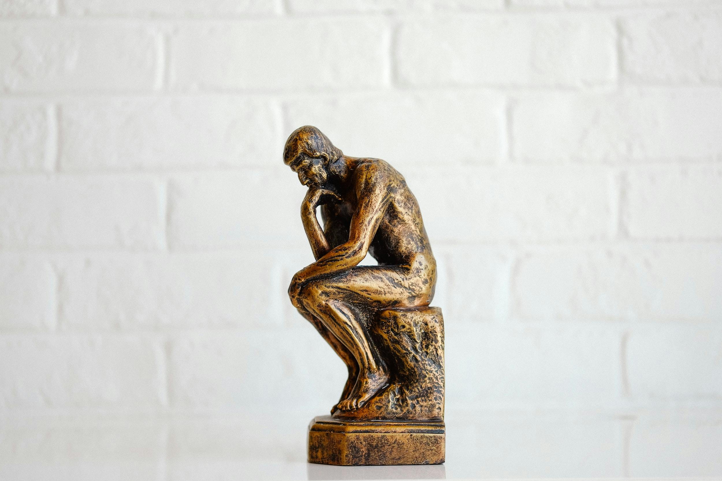 Statuetta di bronzo de "Il Pensatore" su sfondo bianco: Uomo in posizione seduta con una mano poggiata sotto il mento in posizione riflessiva.