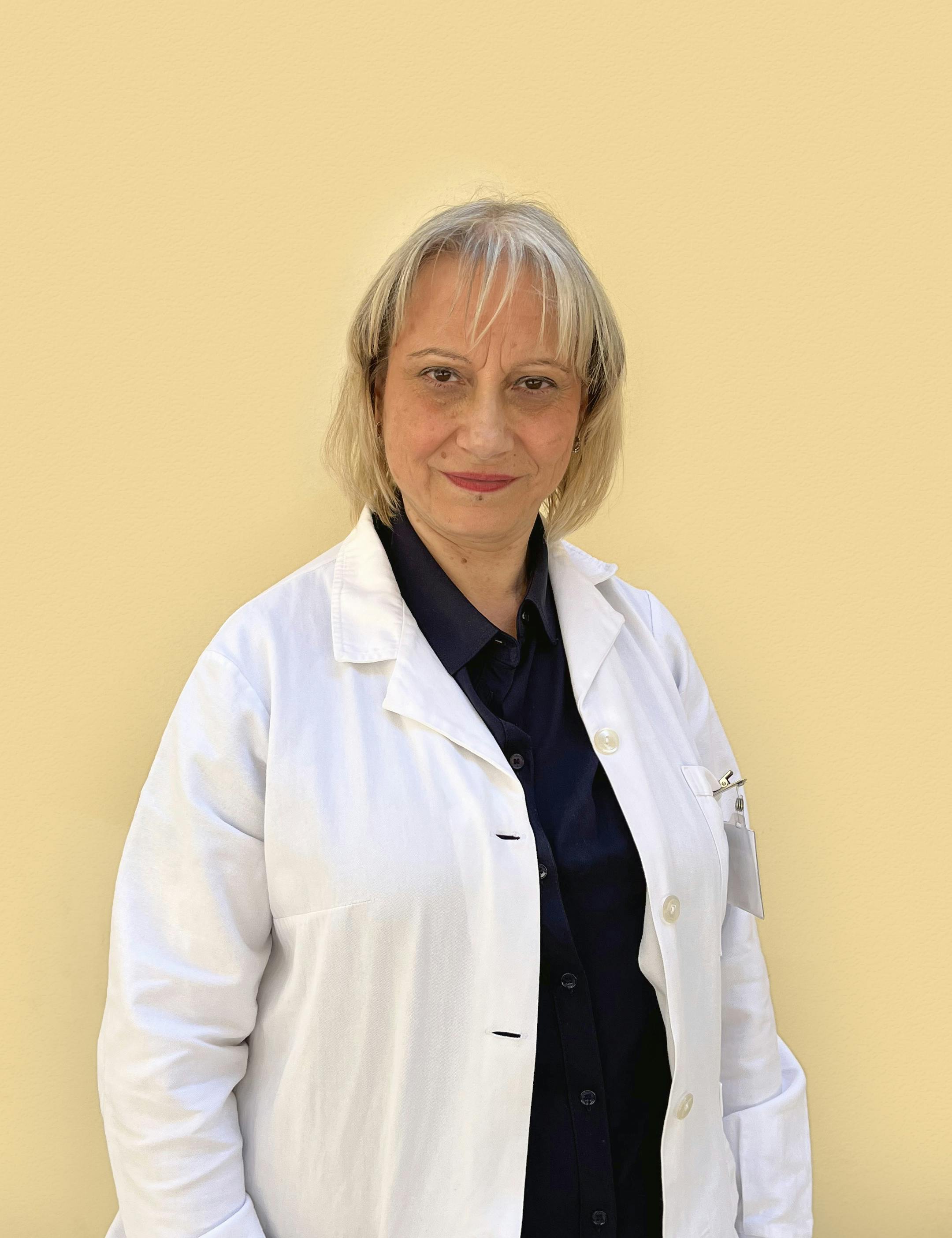 Middle-aged woman with bob blond hair, white doctor's coat and dark blue shirt slightly smiling looking directly at the camera.