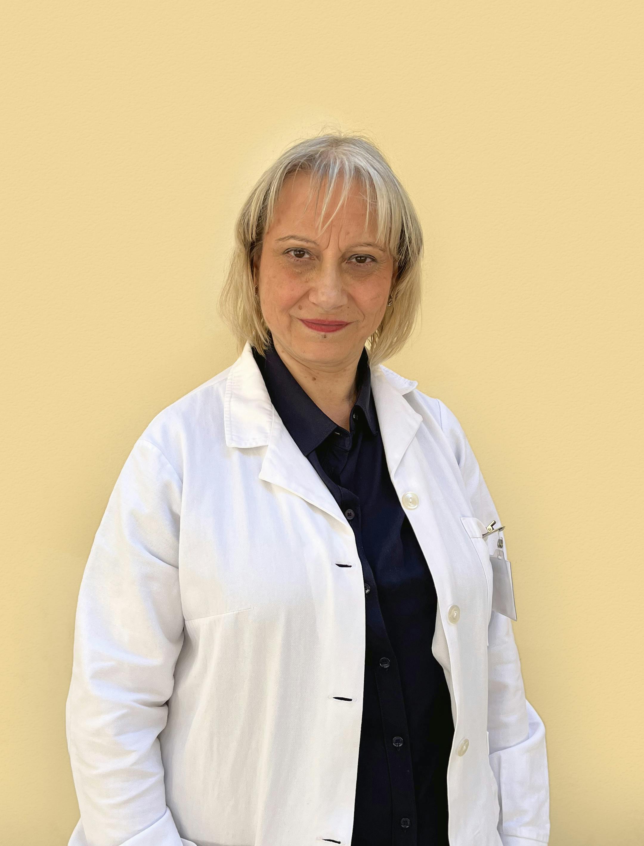 Middle-aged woman with bob blond hair, white doctor's coat and dark blue shirt slightly smiling looking directly at the camera.