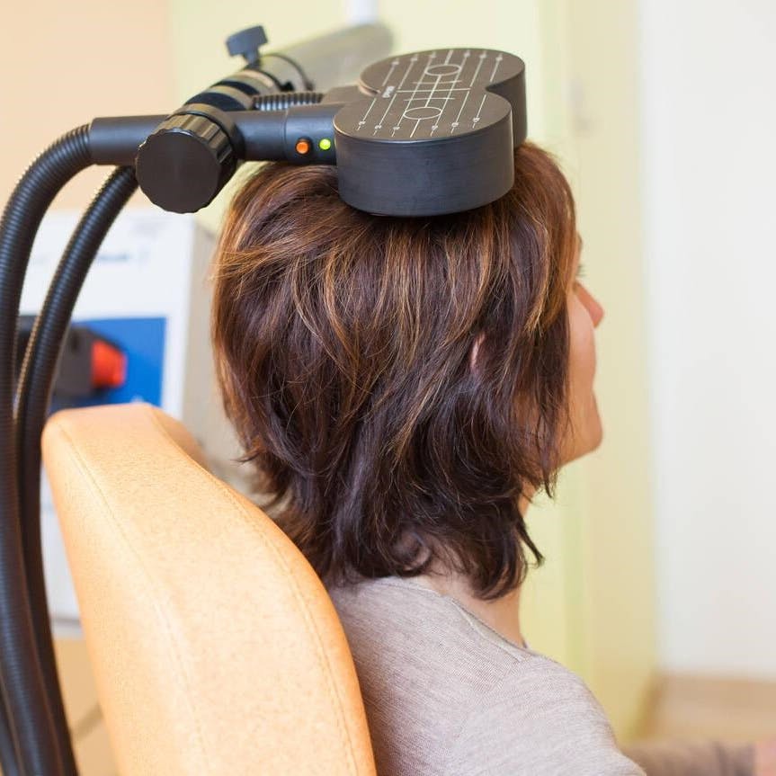 Woman sitting in an orange chair with a magnetic coil used for Transcranial Magnetic Stimulation resting on her head.