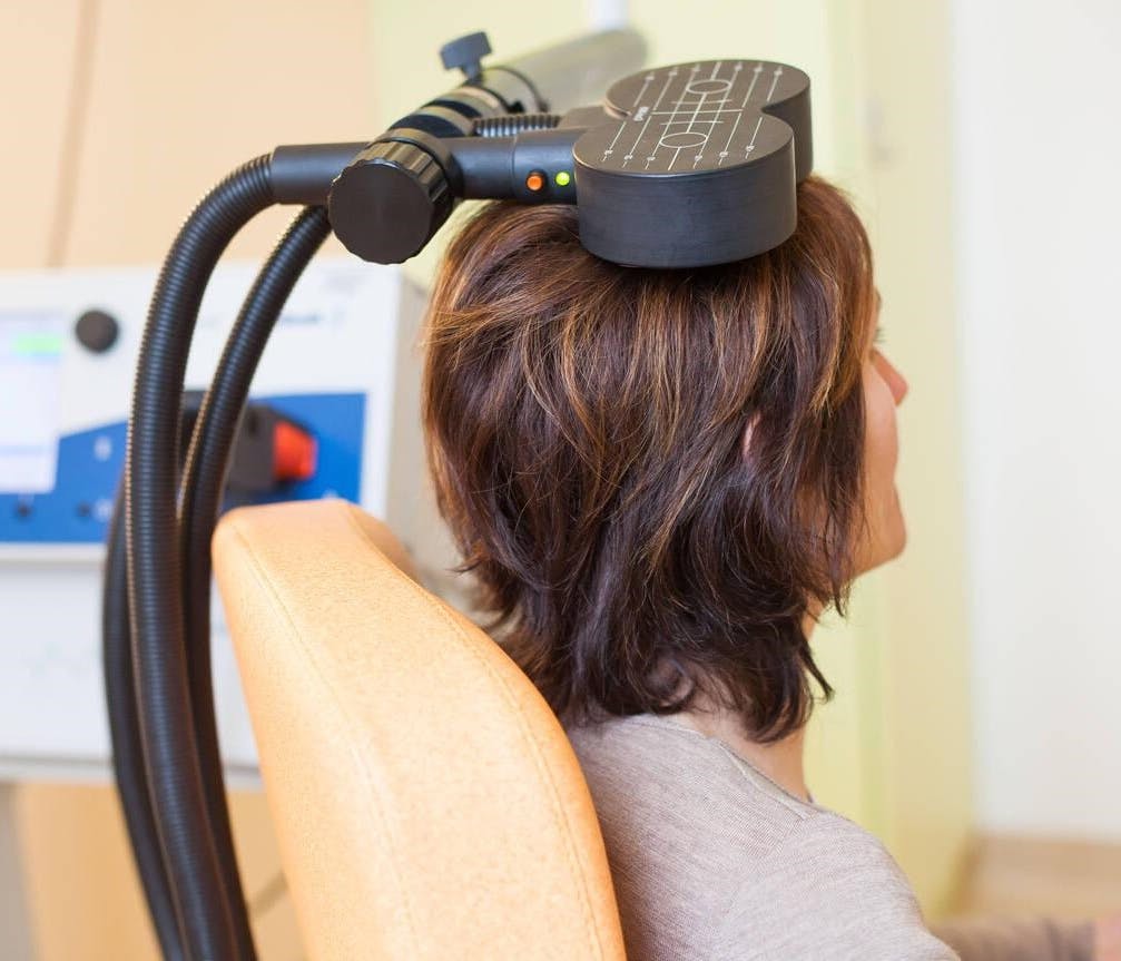 Woman sitting in an orange chair with a magnetic coil used for Transcranial Magnetic Stimulation resting on her head.