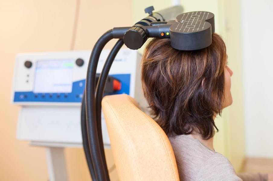 Woman sitting in an orange chair with a magnetic coil used for Transcranial Magnetic Stimulation resting on her head.