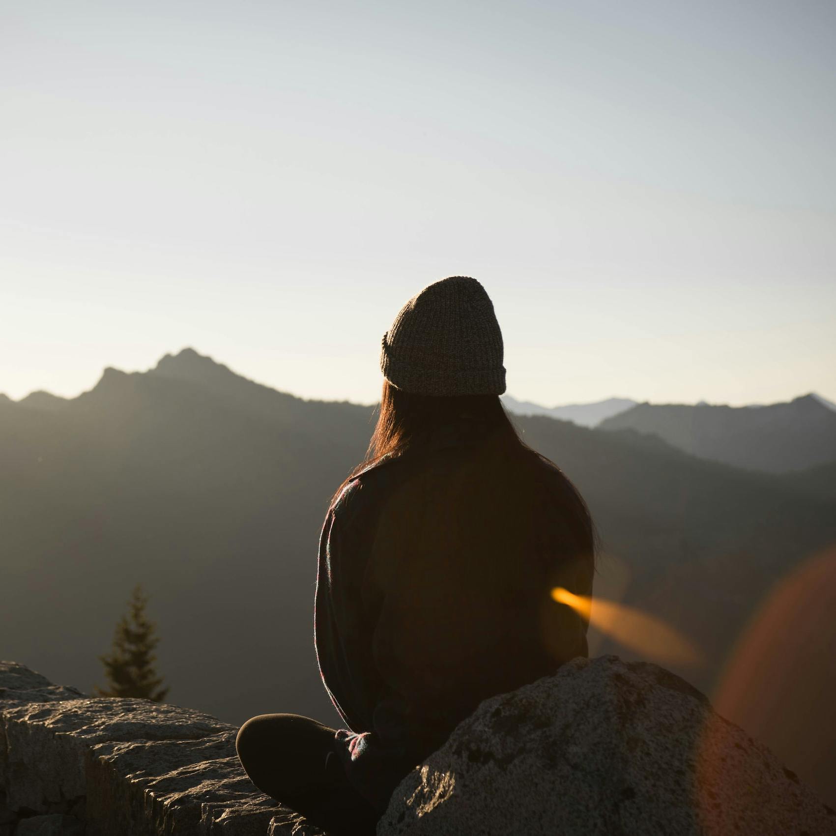 Fotografia di una donna vista di spalle seduta su un terreno roccioso che contempla un paesaggio montano con il sole che la illumina frontalmente.