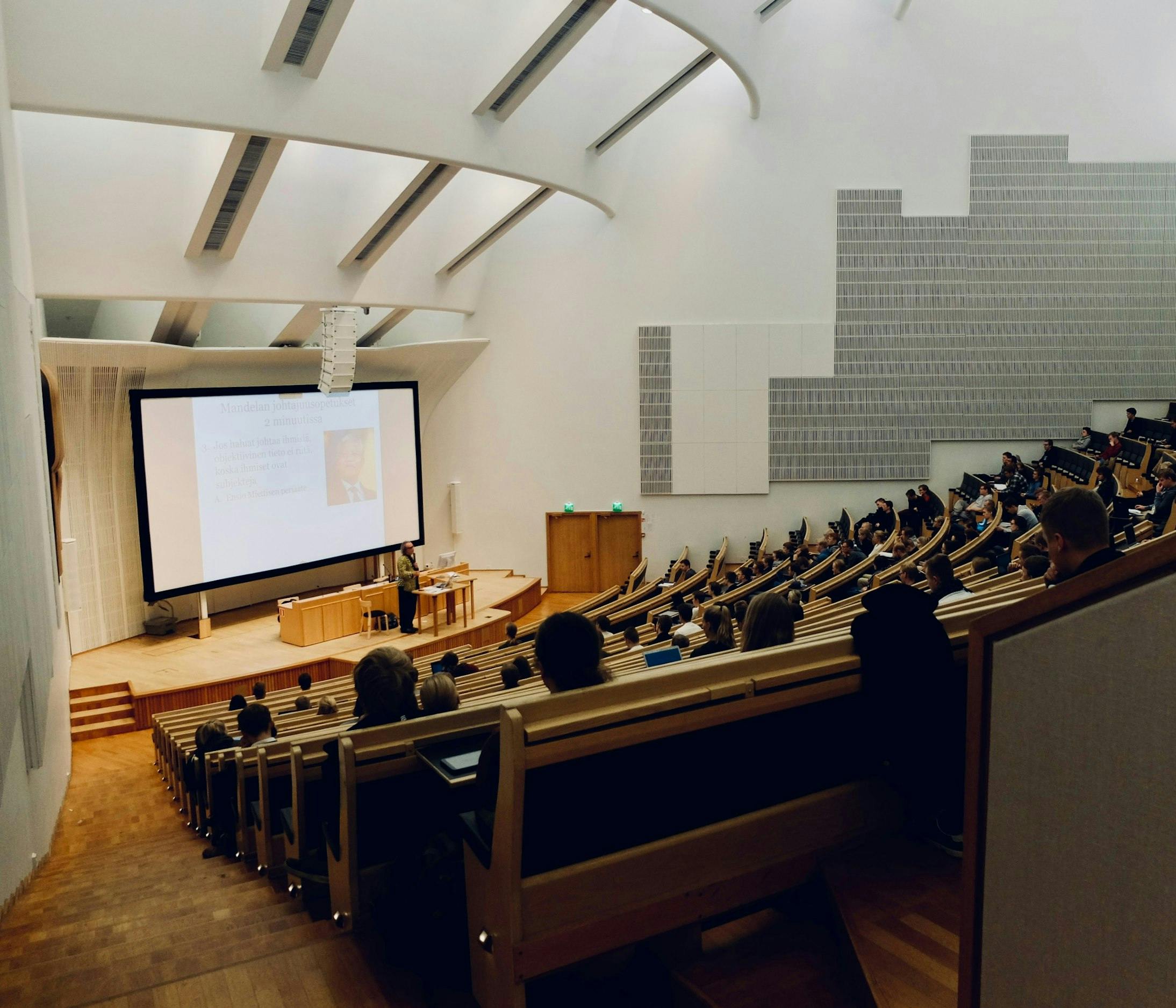 Persone sedute in una sala da congresso ad anfiteatro ad ascoltare una lezione di un professore.