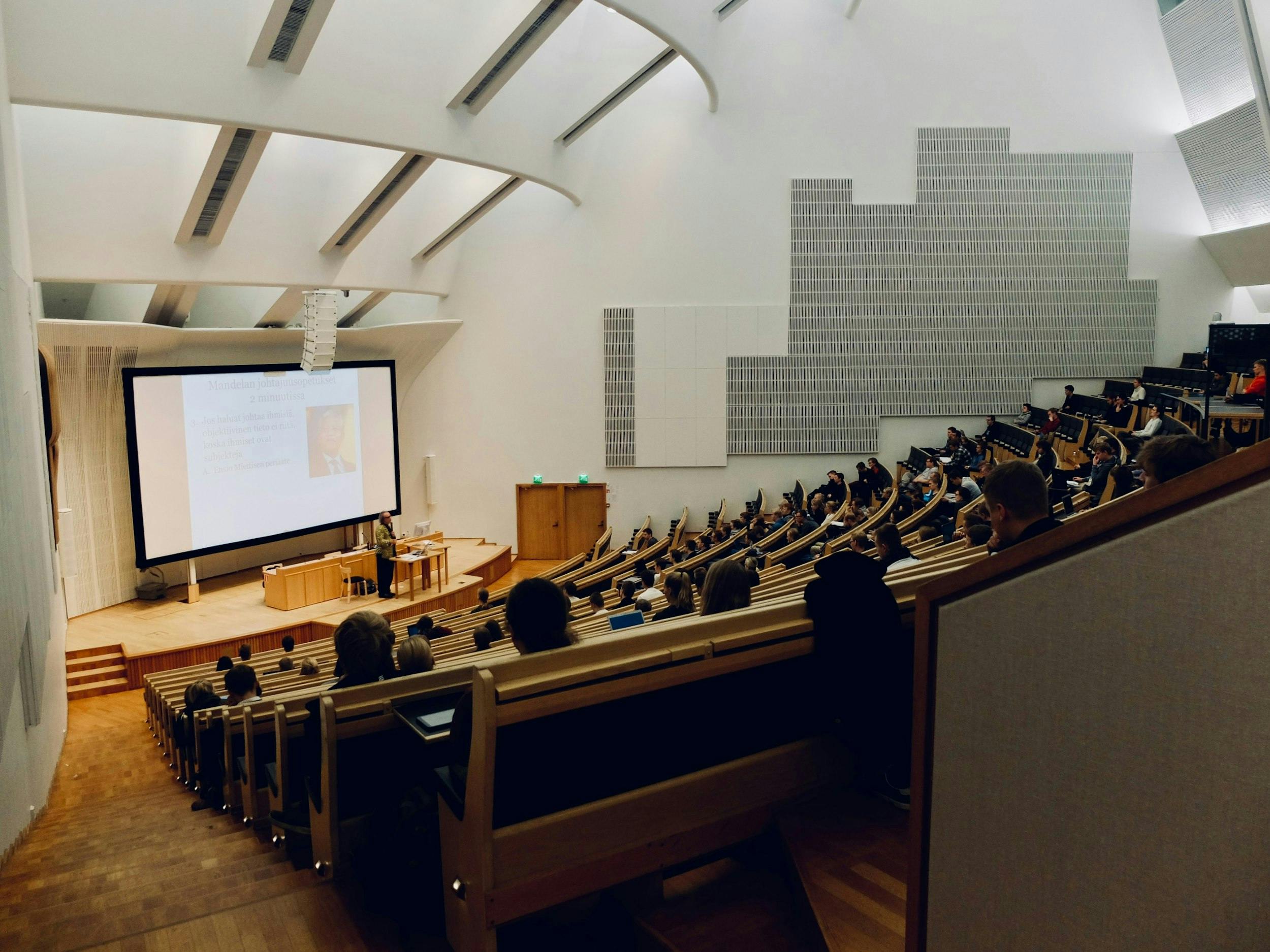 Persone sedute in una sala da congresso ad anfiteatro ad ascoltare una lezione di un professore.