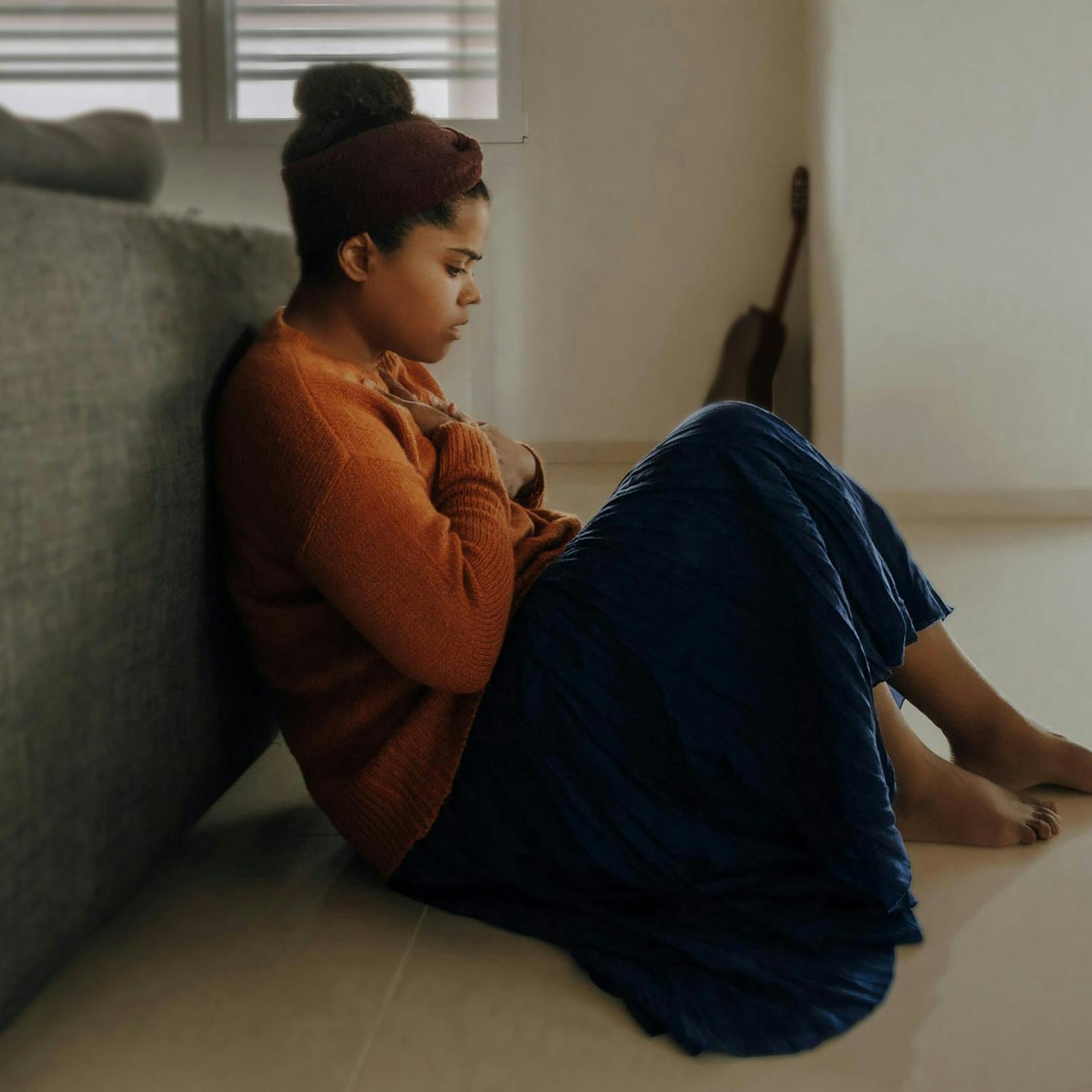 A dark-skinned woman sitting on the floor with her back leaning against a couch bringing her hand to her chest as a sign of concern.