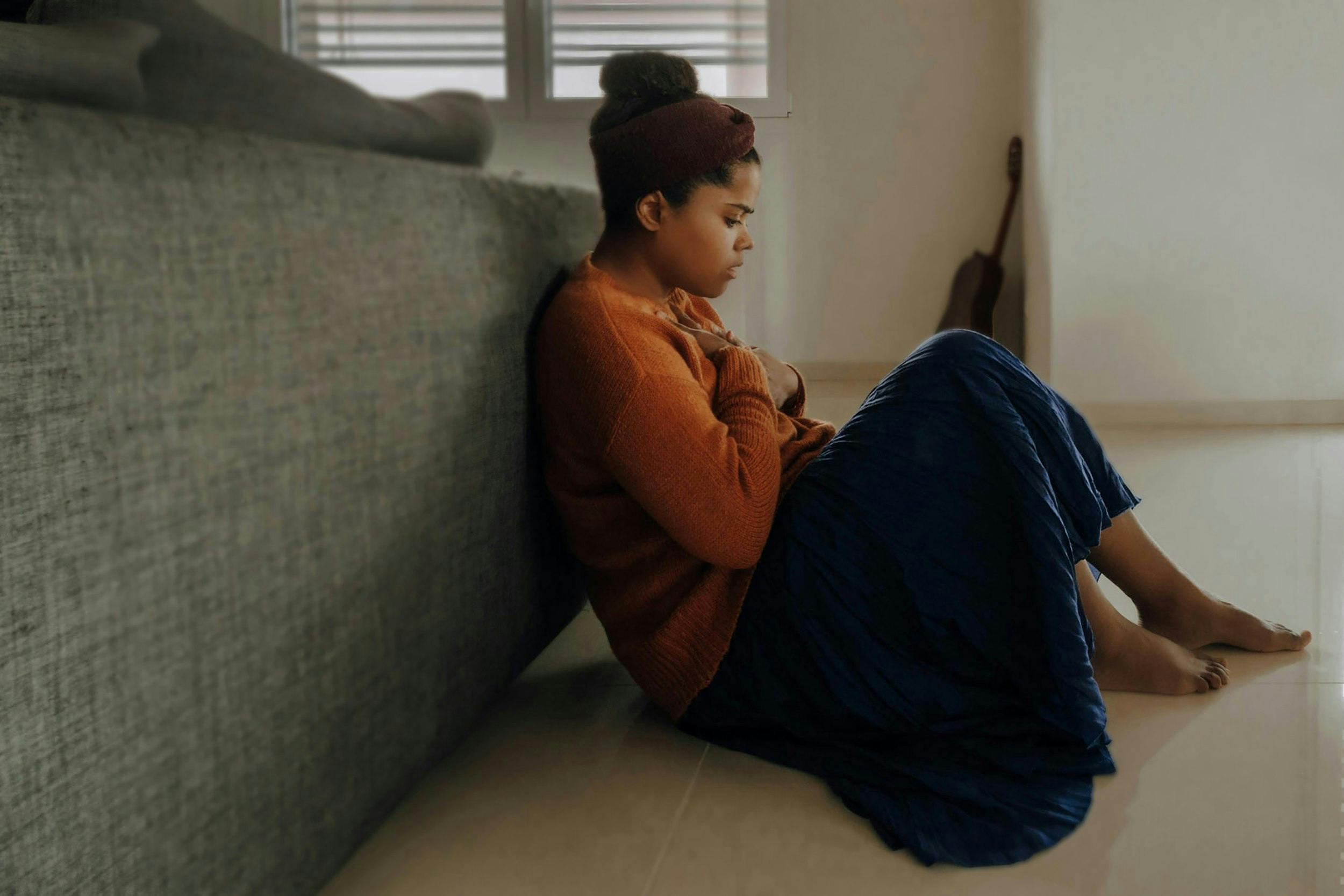 A dark-skinned woman sitting on the floor with her back leaning against a couch bringing her hand to her chest as a sign of concern.