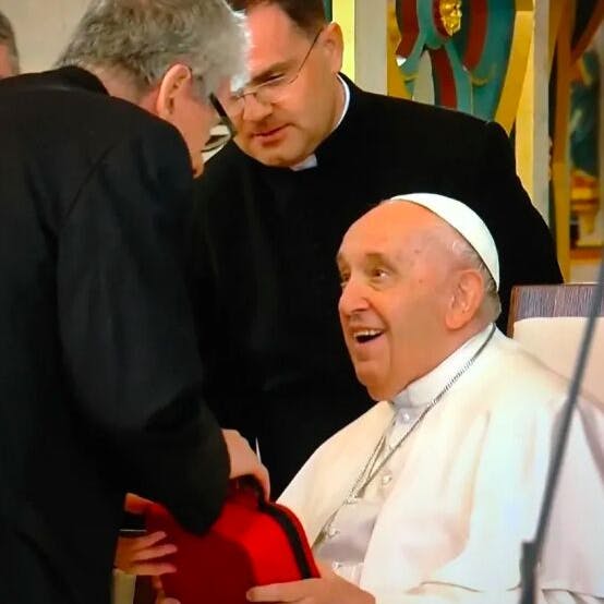 Pope Francis sitting on a chair while receiving from Adam Mester as a gift a Photo-Bio-Modulation device contained inside a red case.