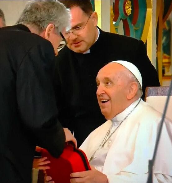Pope Francis sitting on a chair while receiving from Adam Mester as a gift a Photo-Bio-Modulation device contained inside a red case.