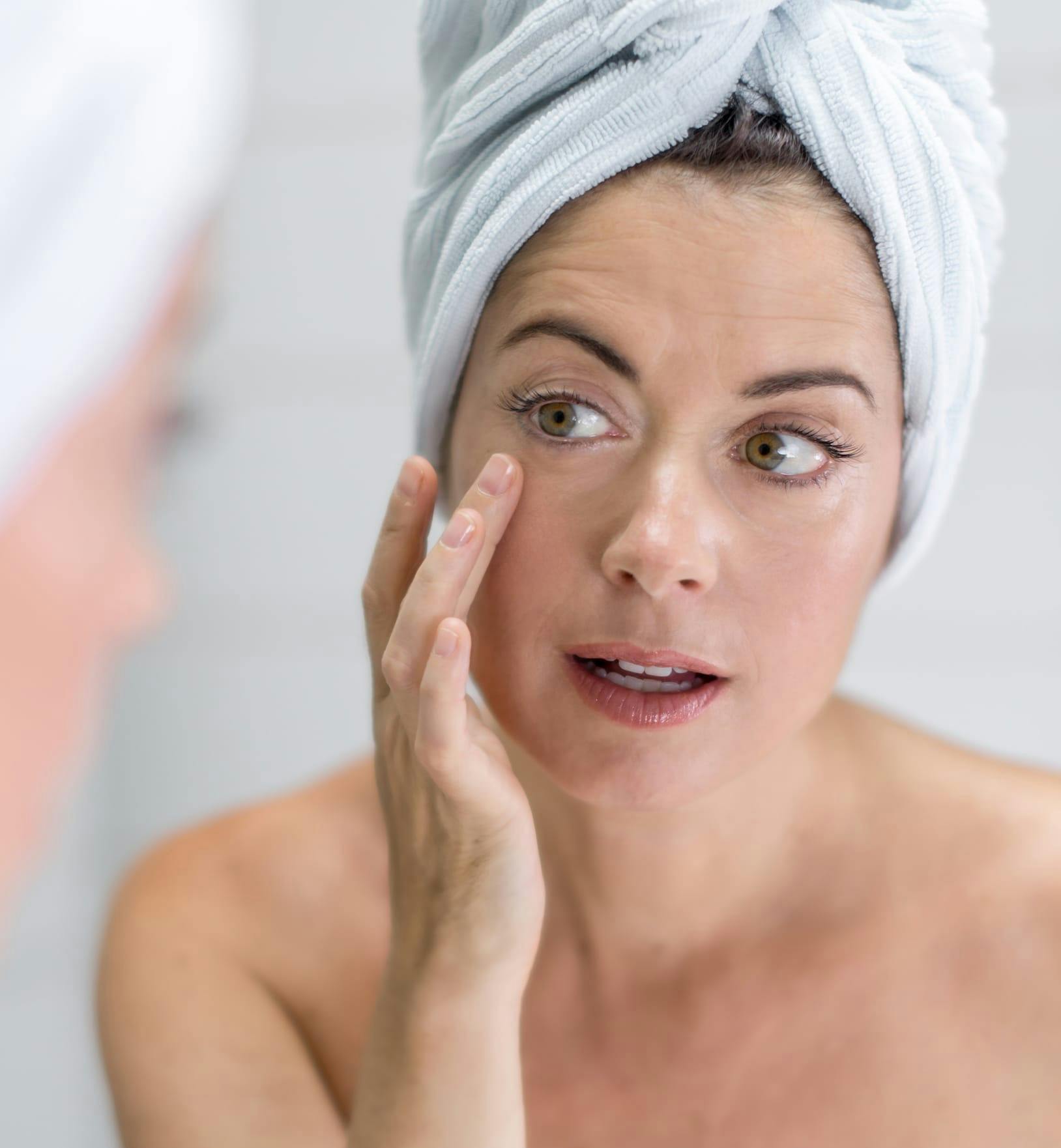 Woman with her hair in a towel on top of her head looking at herself in the mirror