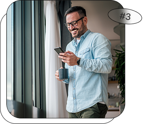 homme près d'une fenêtre qui sourit en regardant son téléphone et avec une tasse à la main
