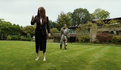still from the movie Bugonia featuring a man with a mask following a woman on a grassy lawn