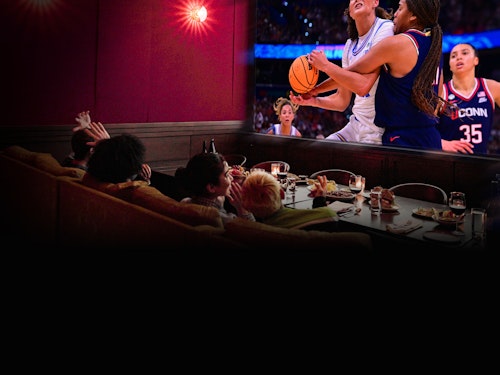 A group of people in a dimly lit restaurant booth reacts excitedly to a women's basketball game shown on a large screen. The atmosphere is lively and engaging.