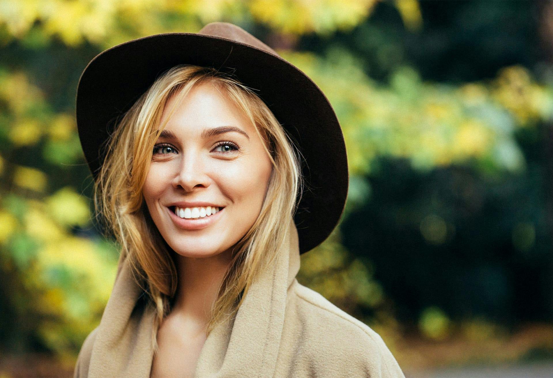 Woman wearing a hat smiling in front of foliage.