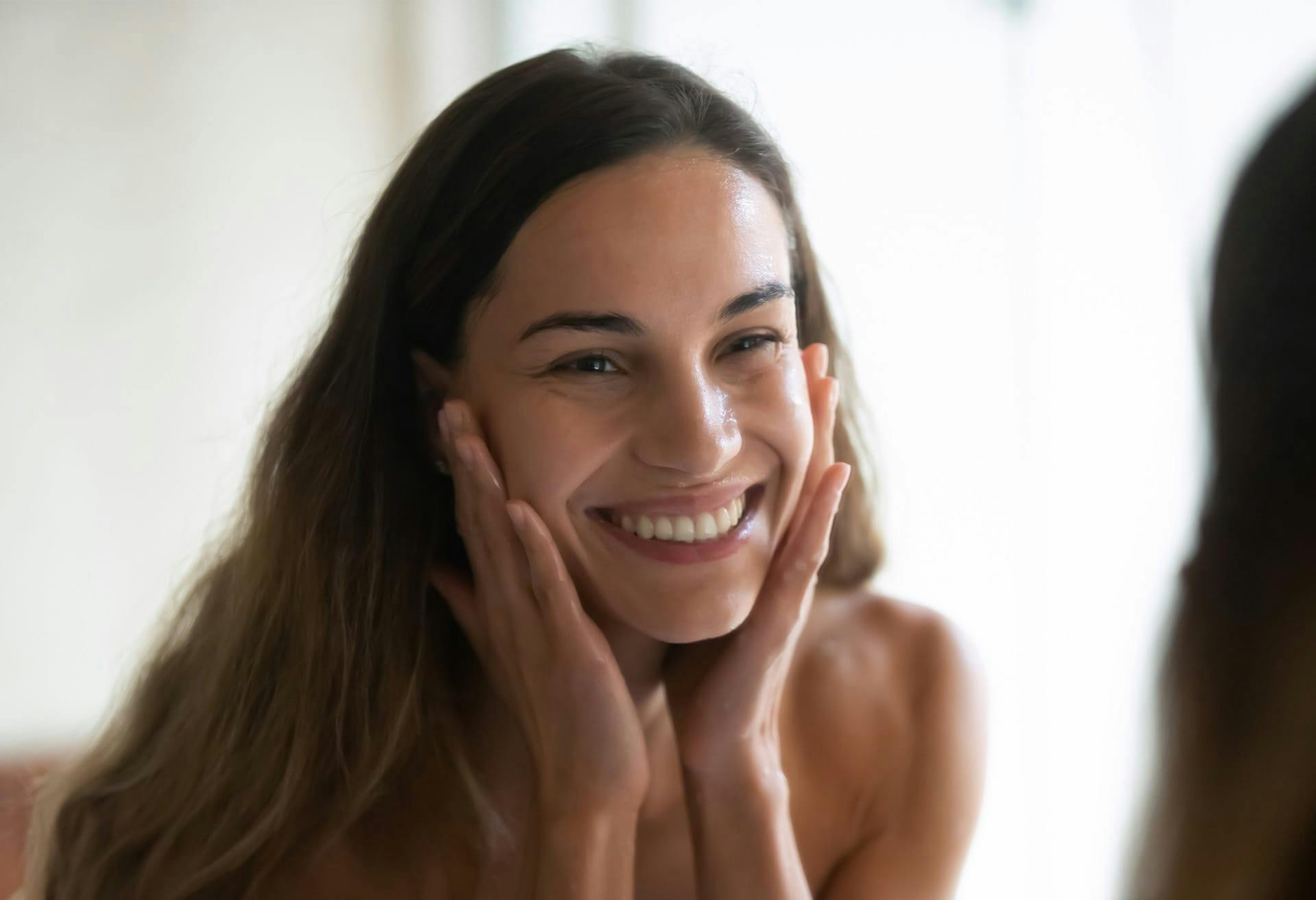 tan brunette model smiling into a mirror with her hands up to her face