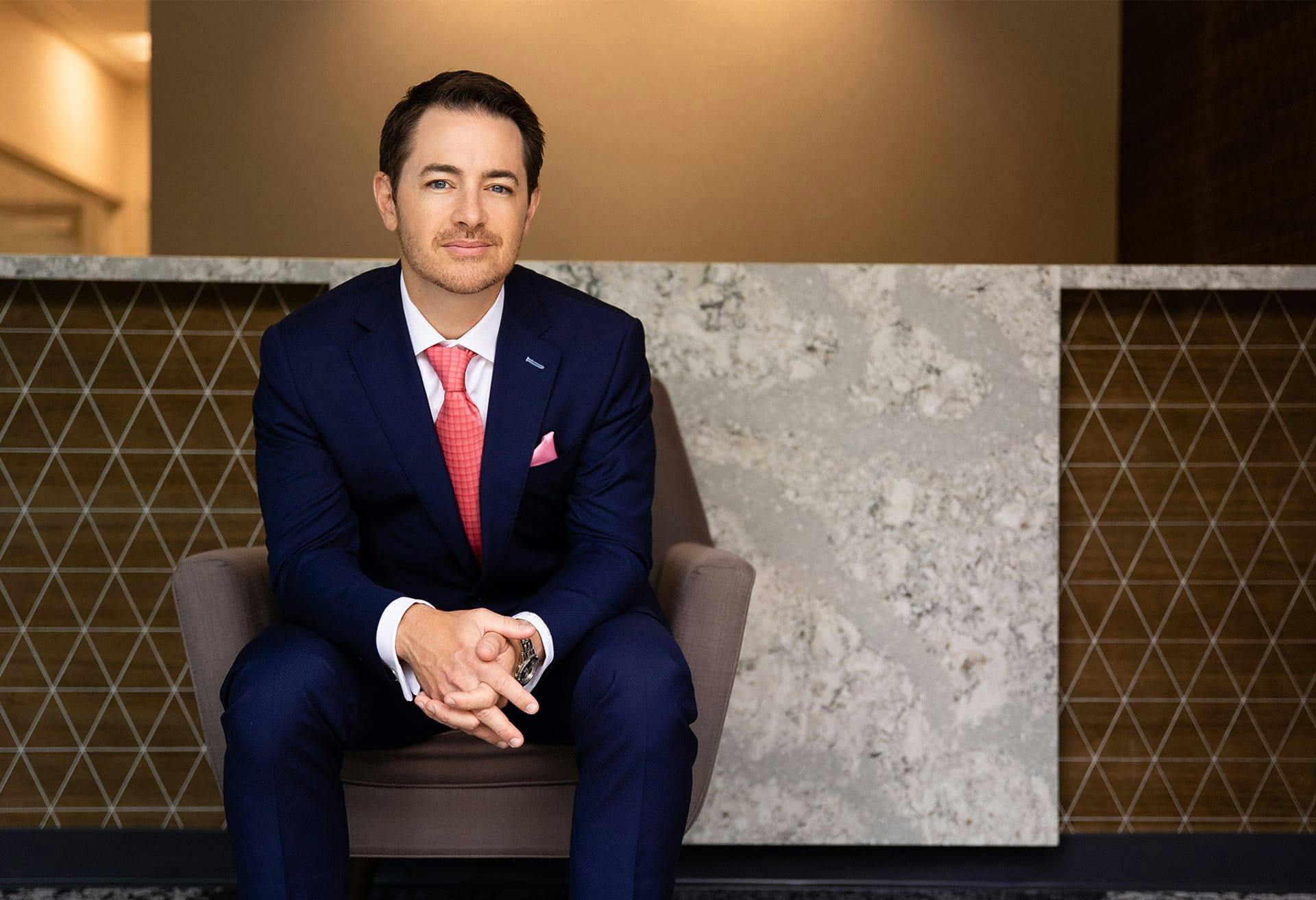 Dr. Robb sitting in a chair in the lobby in a dark navy suit and pink tie