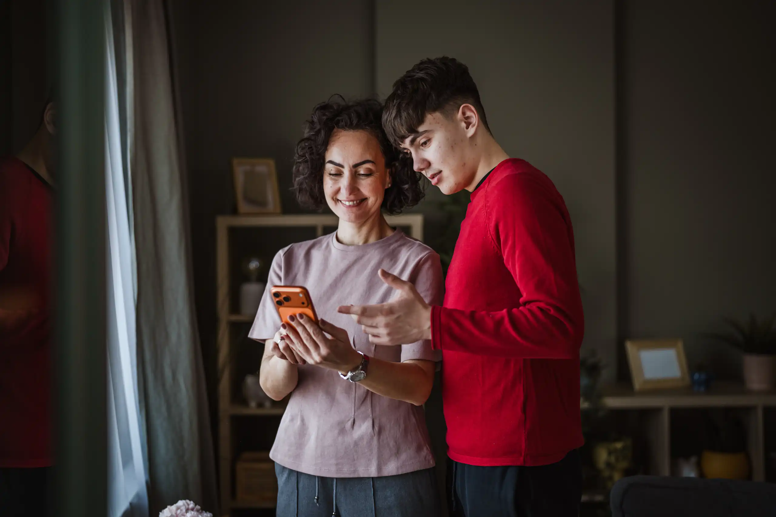 Everyday moments, like checking a message together, can open the door to bigger conversations. Mother and teenage son smiling together while looking at a phone at home