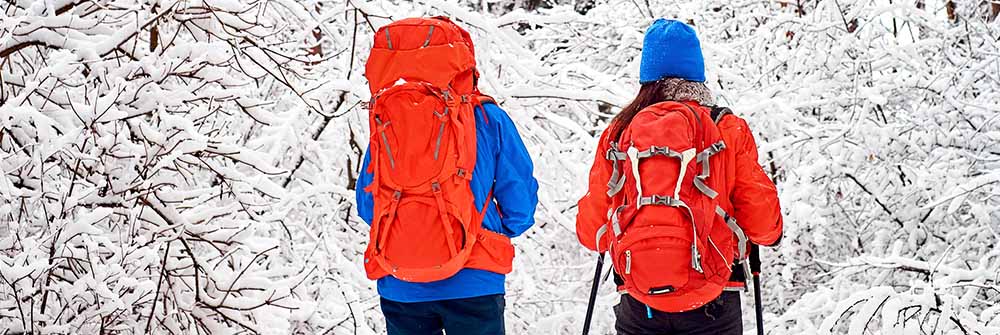 Två personer med röda vandringsryggsäckar vandrar genom en snötäckt skog, utrustade för vintervandring och friluftsliv