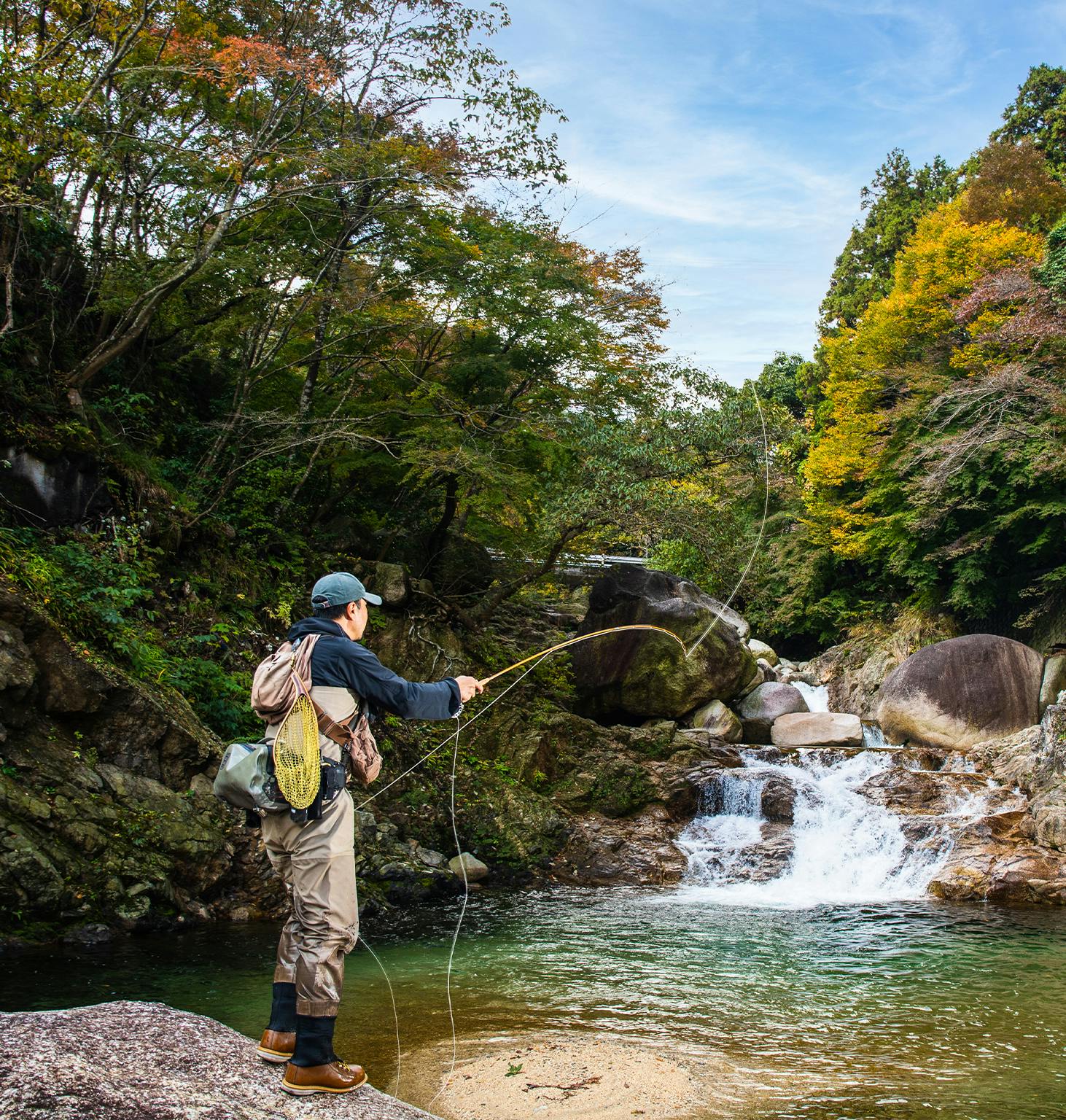 man fly fishing in a stream in the woods with a waterfall