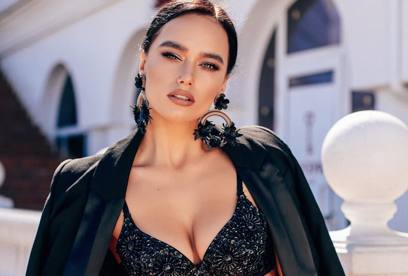 a woman with black hair wearing a black blazer and bra, with large black floral earrings