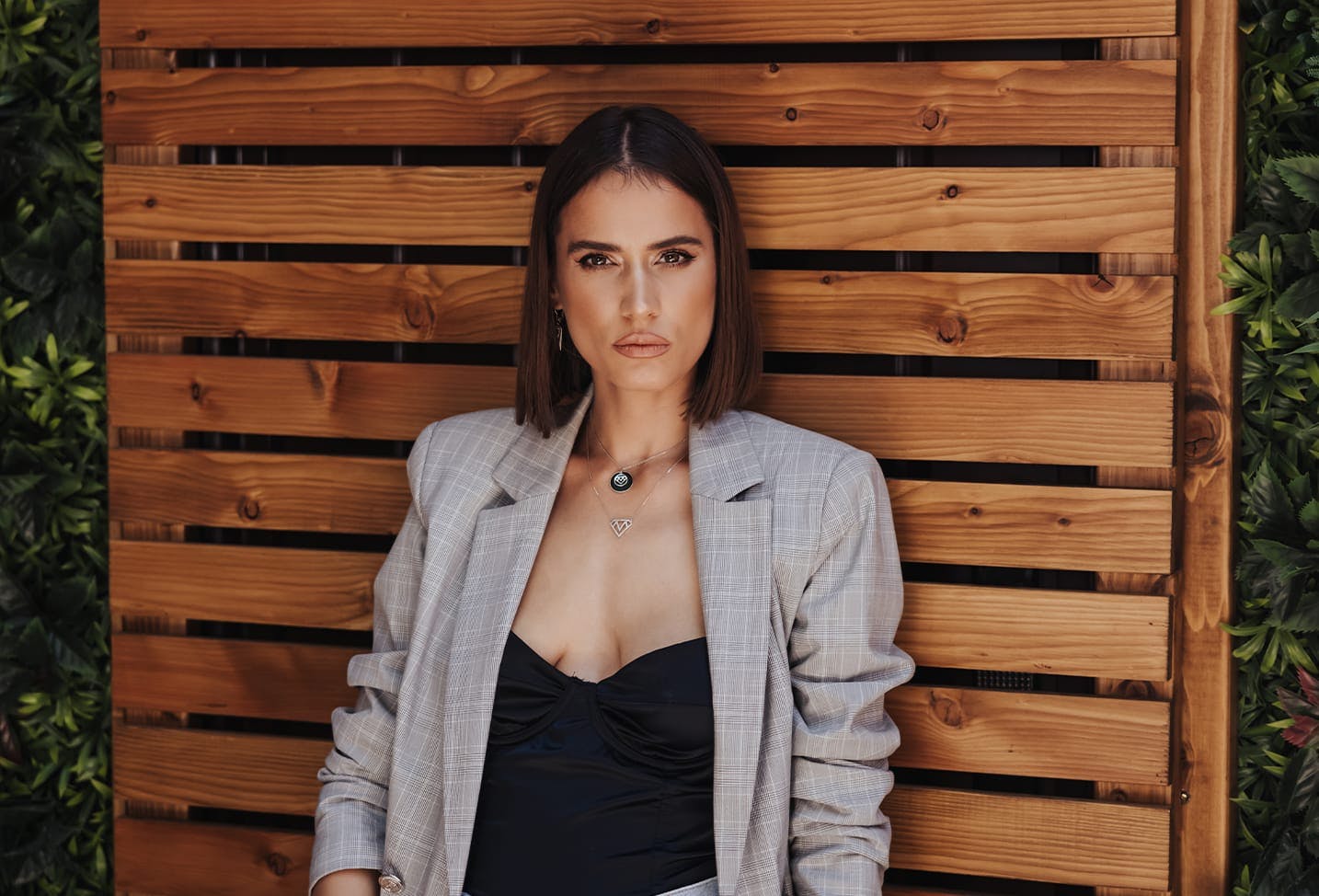a woman with short dark brunette hair leaning against a wooden fence, wearing a grey blazer and black top