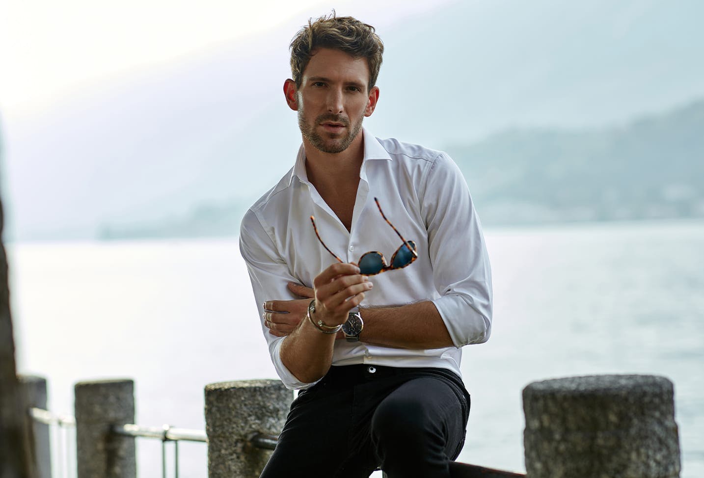 a man sitting by the pier wearing a white shirt, black pants, and holding sunglasses