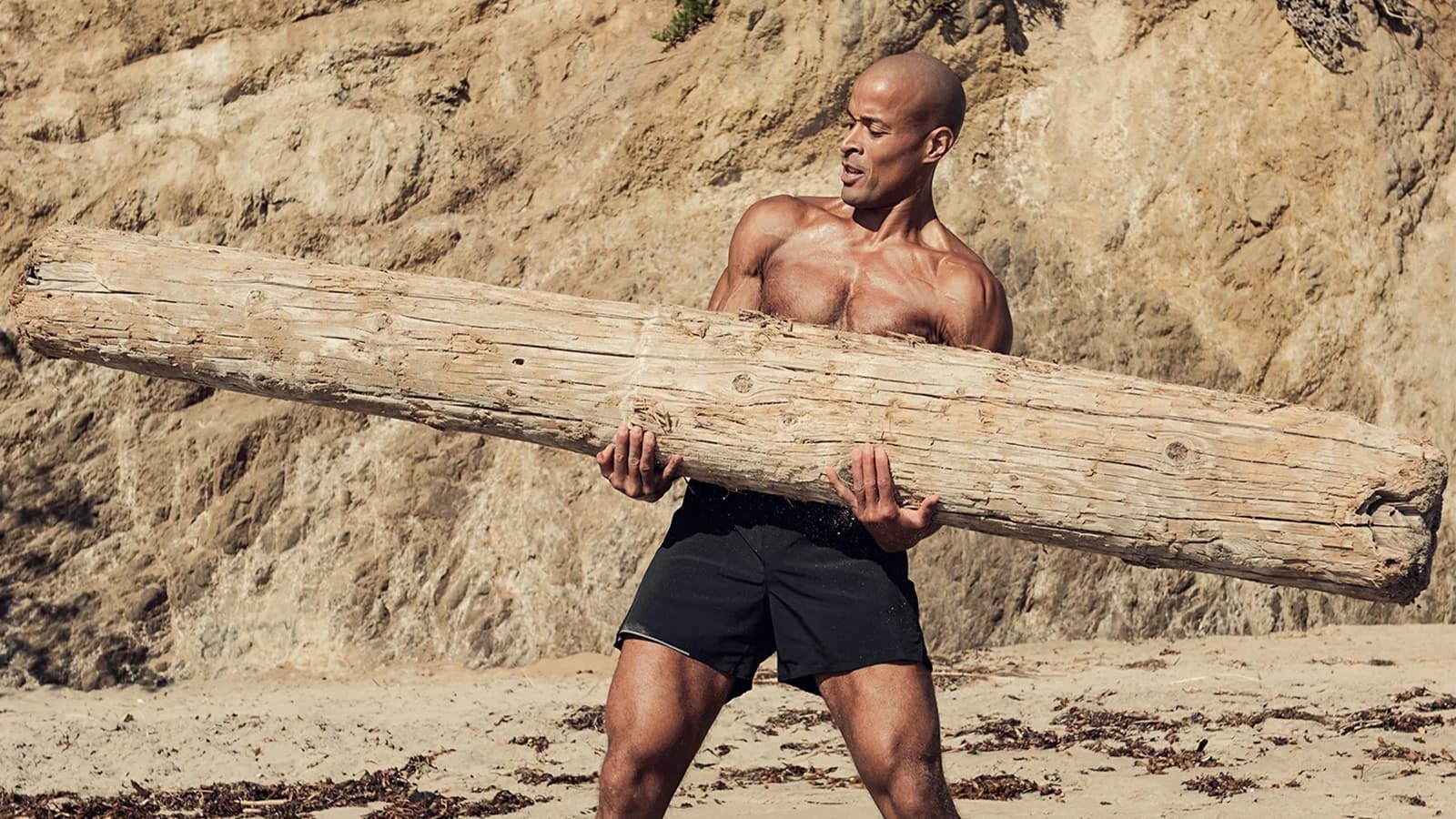 David Goggins mit einem Baumstamm in der Hand an einem Strand mit Felsen im Hintergrund.