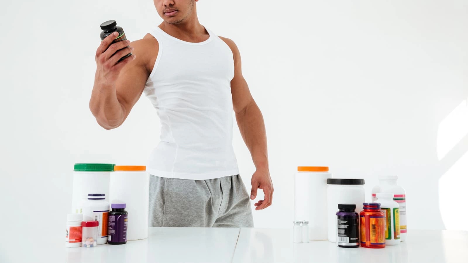 Bild von einem muskulösen Mann hinter einem Tisch voller Supplements.