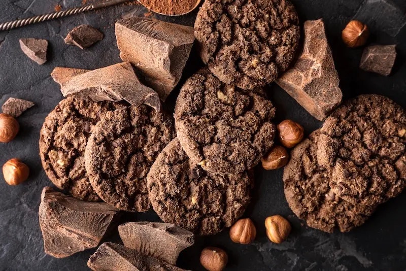 Mehrere Cookies, Schokolade und Nüsse auf dunklem Grund.