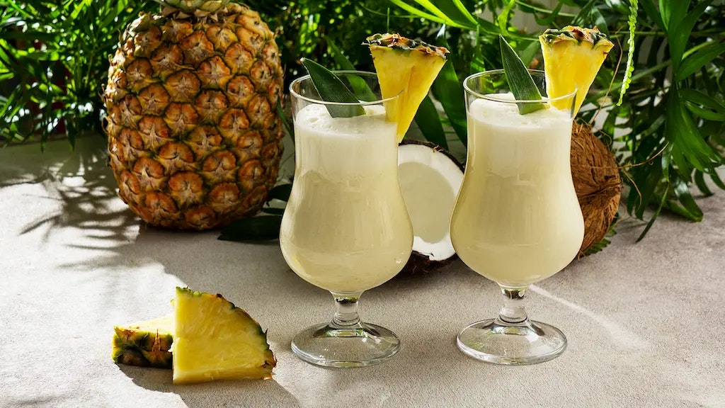 Ein Bild, welches zwei Gläser mit Pina Colada neben einer Ananas und einer Kokosnuss zeigt.