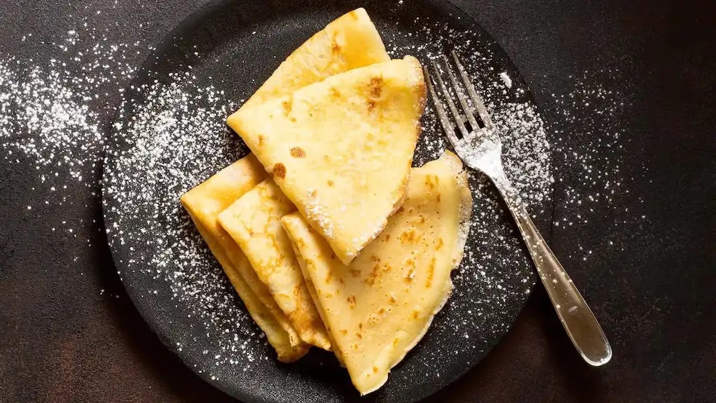 Pfannkuchen mit Puderzucker und eine Gabel auf einem schwarzen Teller.