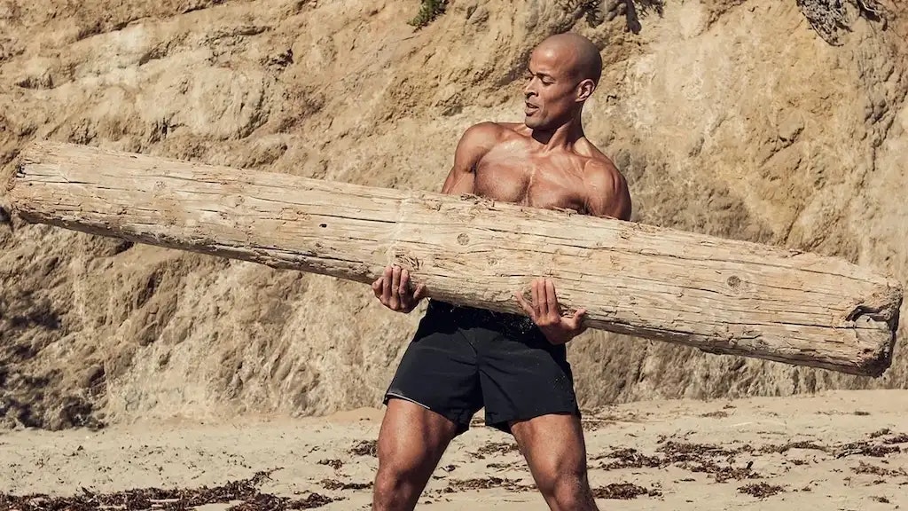 David Goggins mit einem Baumstamm in der Hand an einem Strand mit Felsen im Hintergrund.
