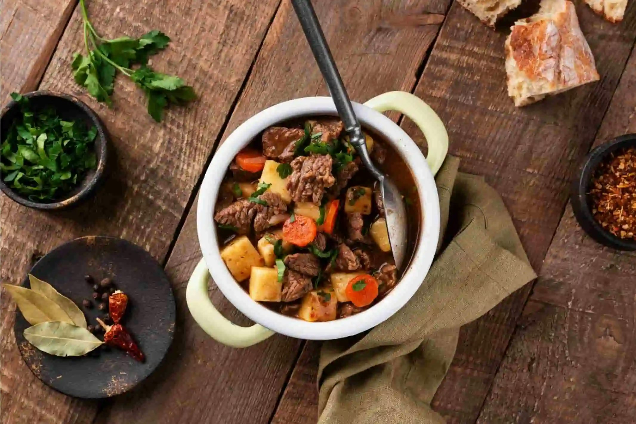 Ein Topf mit klassischen Gulasch. Auf einem Tiasdch, danaben Gewürze, Kräuter und Chili.