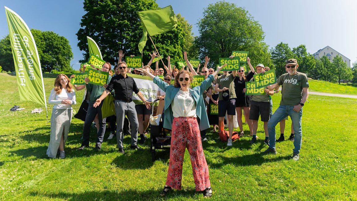 Miljøpartiet De Grønne team family photo on a green lawn, holding flags with their party logo and looking smiling at the camera