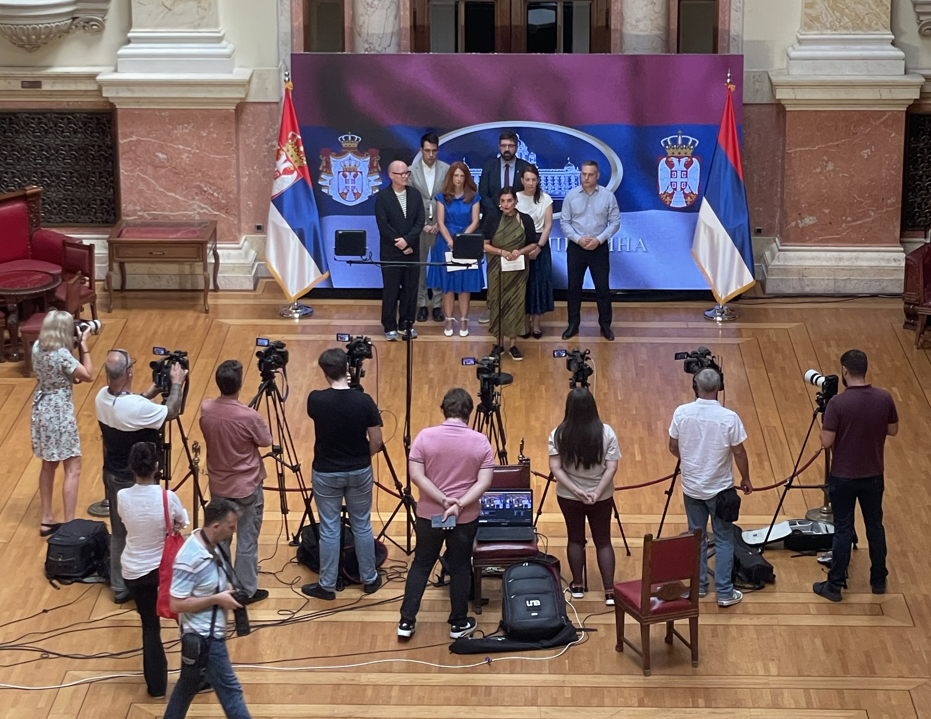 EGP Committee member and co-chair with co-presidents of the Serbian Green-Left Front, standing in front of a Serbian flag, facing journalists with cameras during their press conference.