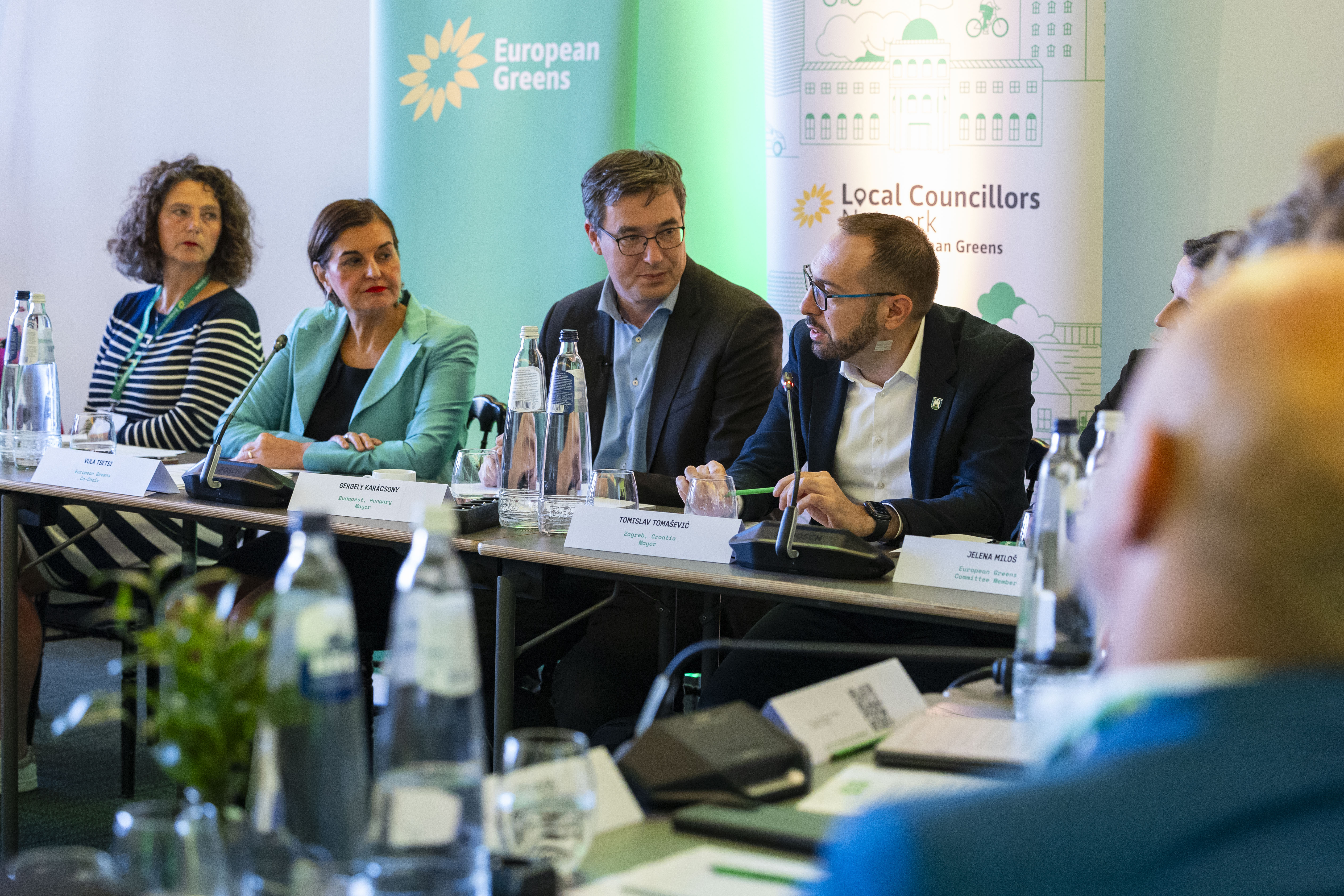 European Greens meeting with mayors and EGP Committee members seated around a conference table, including speakers with microphones and banners for the European Greens and Local Councillors Network in the background.