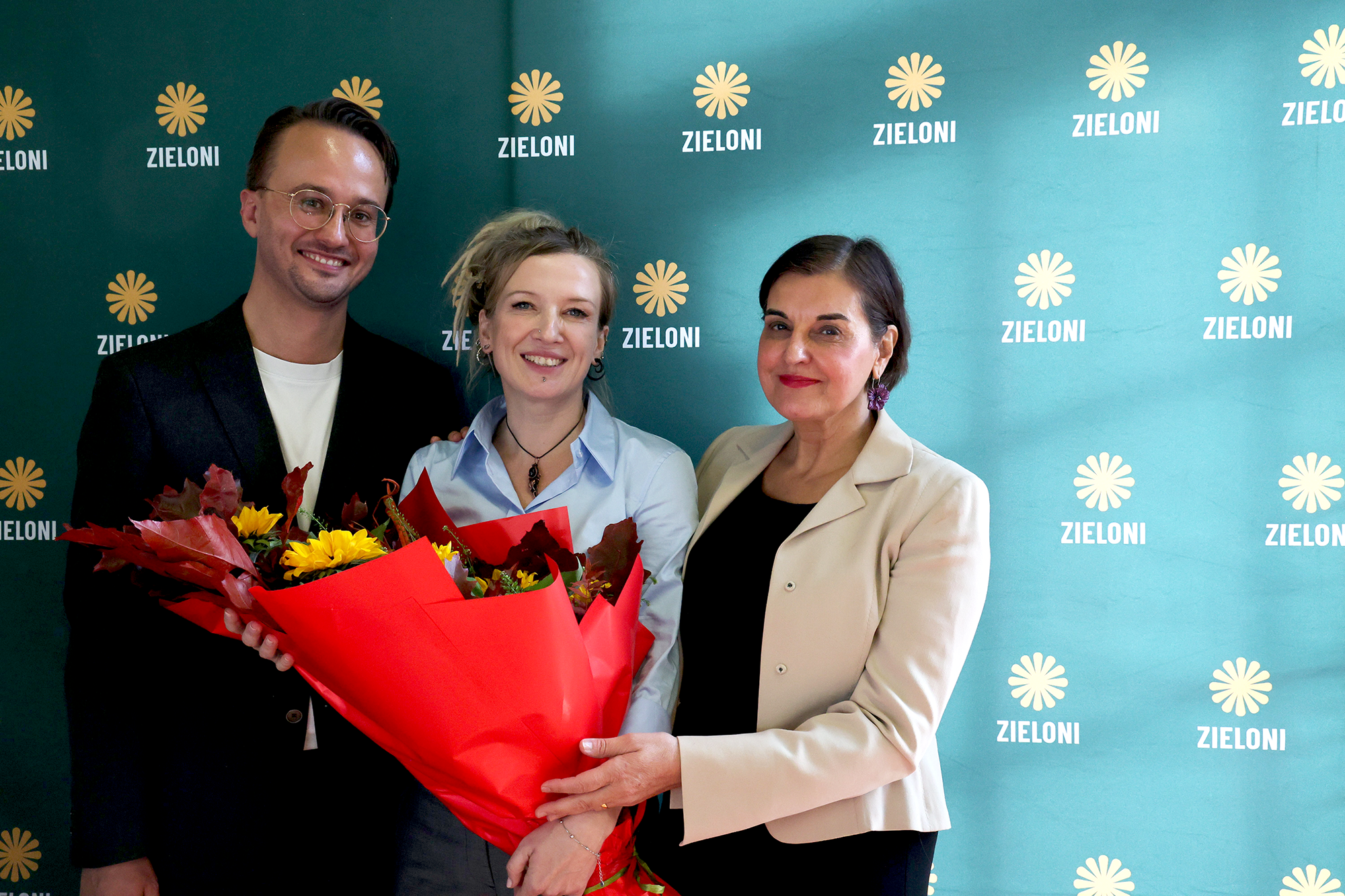 The two new Polish co-chairs and the EGP co-chair are standing in front of a backdrop with Zieloni logo on it, smiling, holding a red and yellow flower bouquet