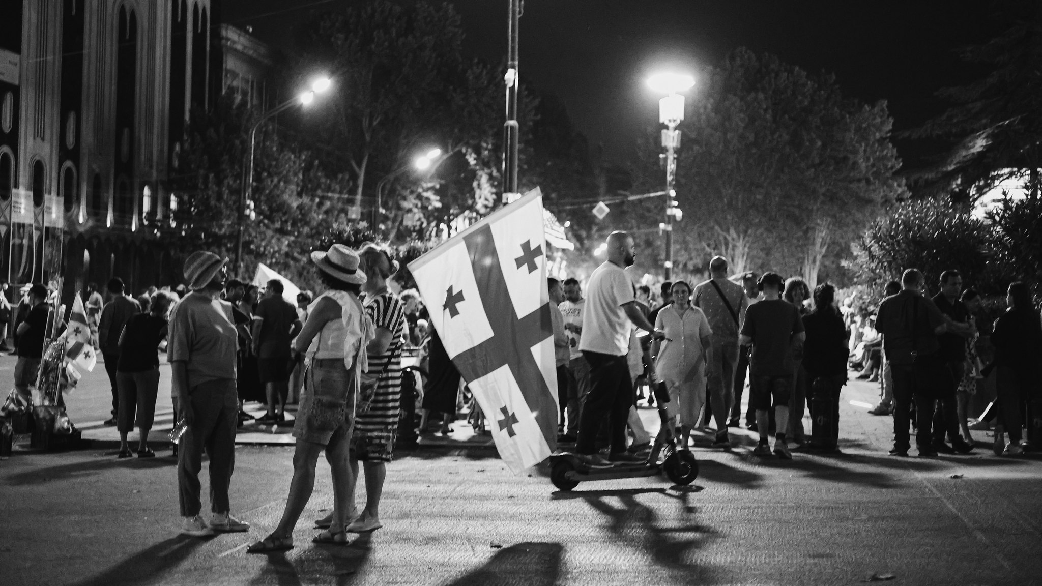 Black and white photo of people standing quietly in a street, one holding a large Georgian flag