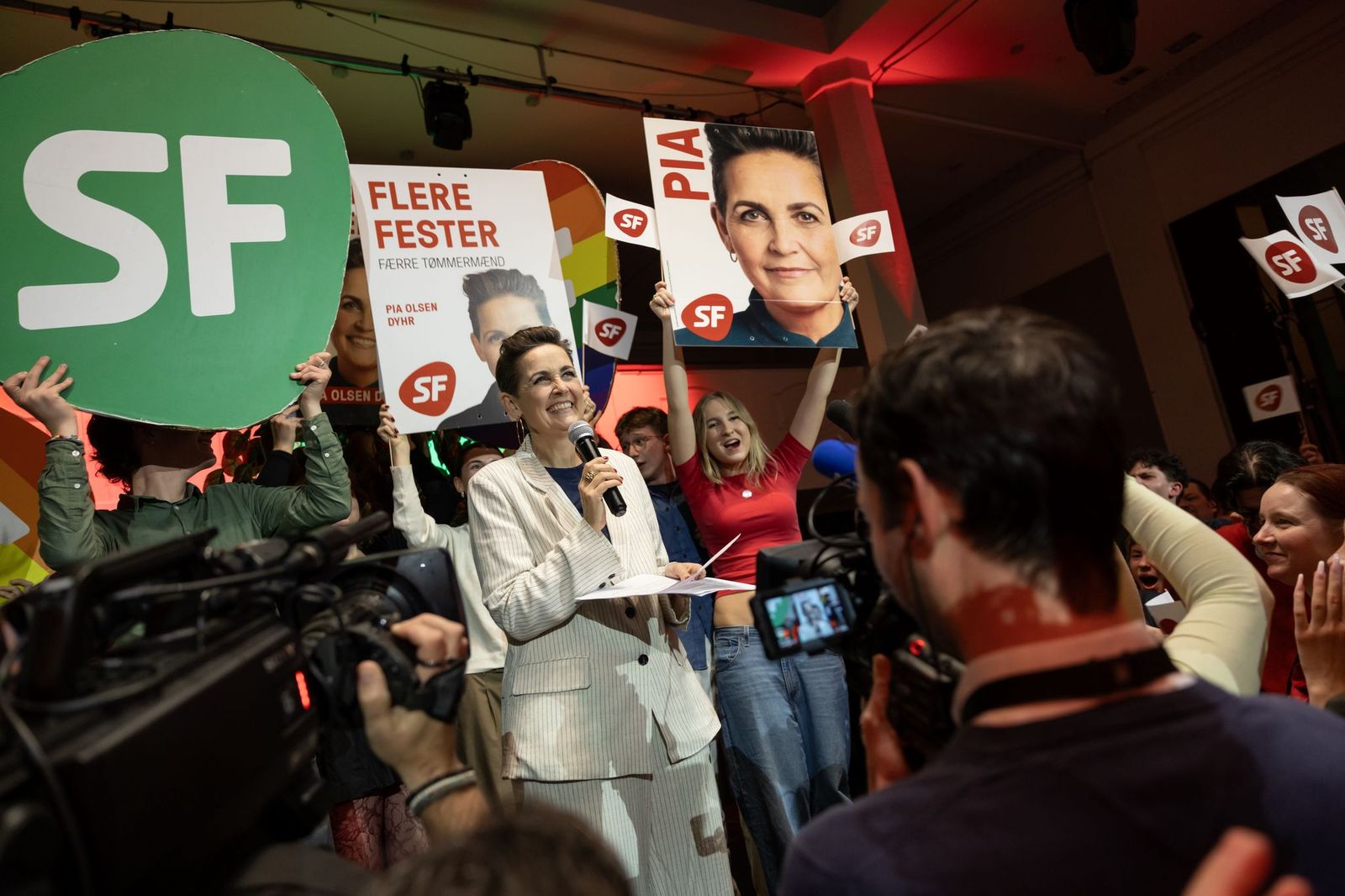 Pia Olsen Dyhr smiling, holding a mic in front of cameras, behind her people holding signs with the SF party logo and Pia's name