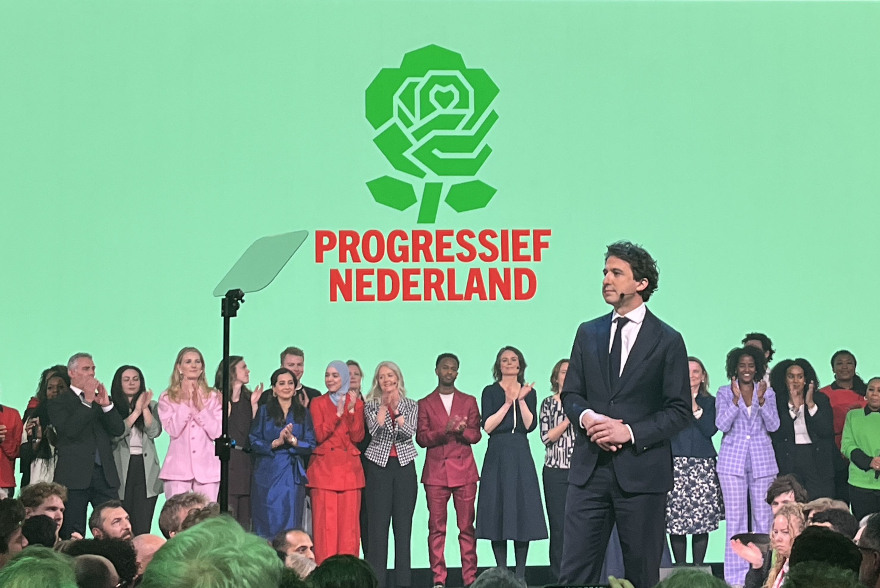 Jesse Klaver on stage in front of a line of personalities from Progressief Nederland Party, with the party logo in the background - a green flower