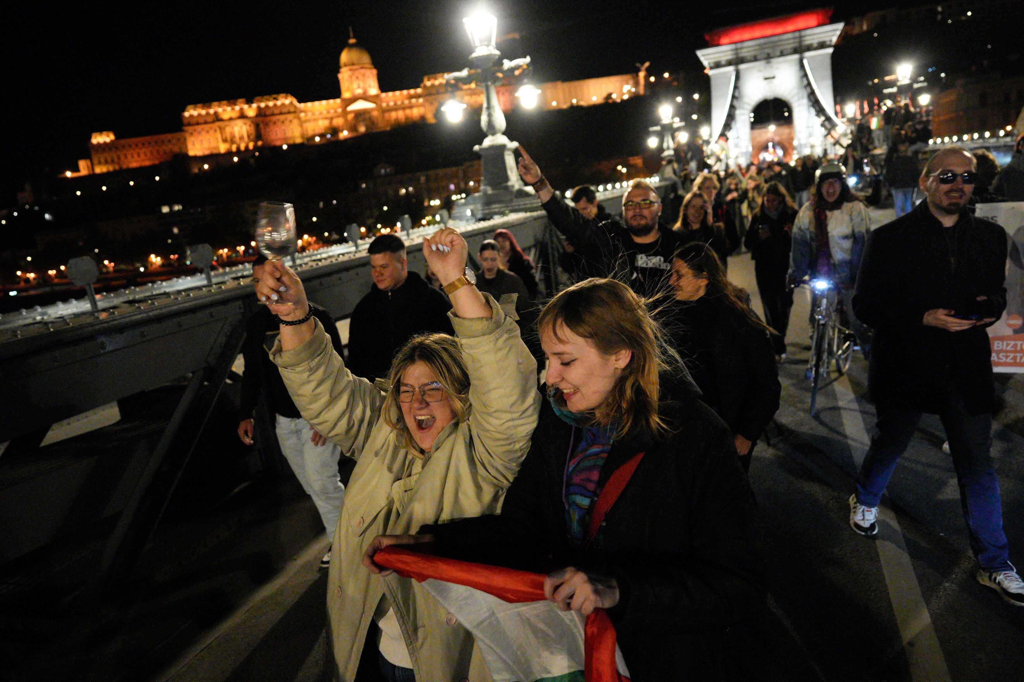 Two women followed by a crowd cheering on a Budapest's bridge at night. They are holding a hungarian flag.
