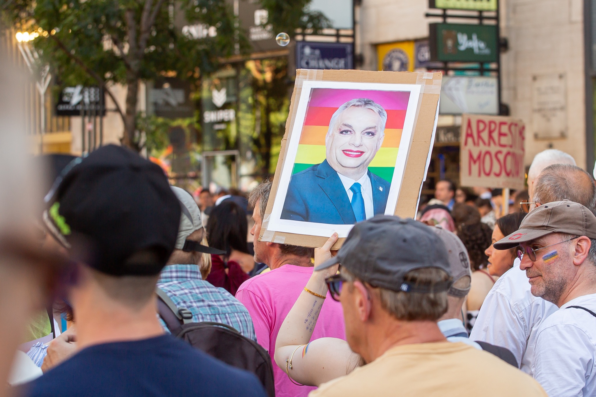 Protesters holding a sign with a portrait of Orban against a rainbow colored background, with fake makeup on, during Budapest Pride 2025