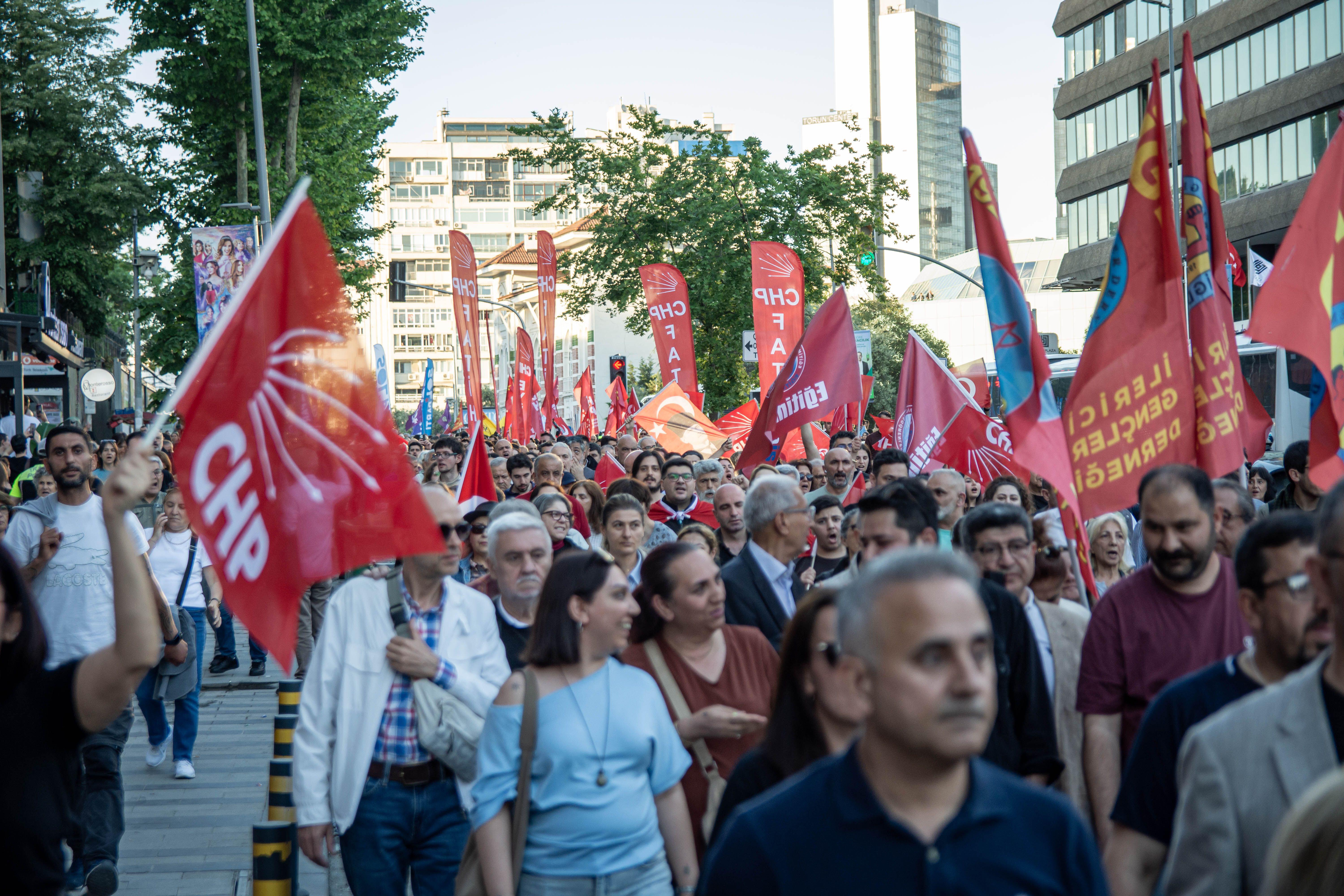 Crowd iin a street in daylight carrying red flags and CHP flags.