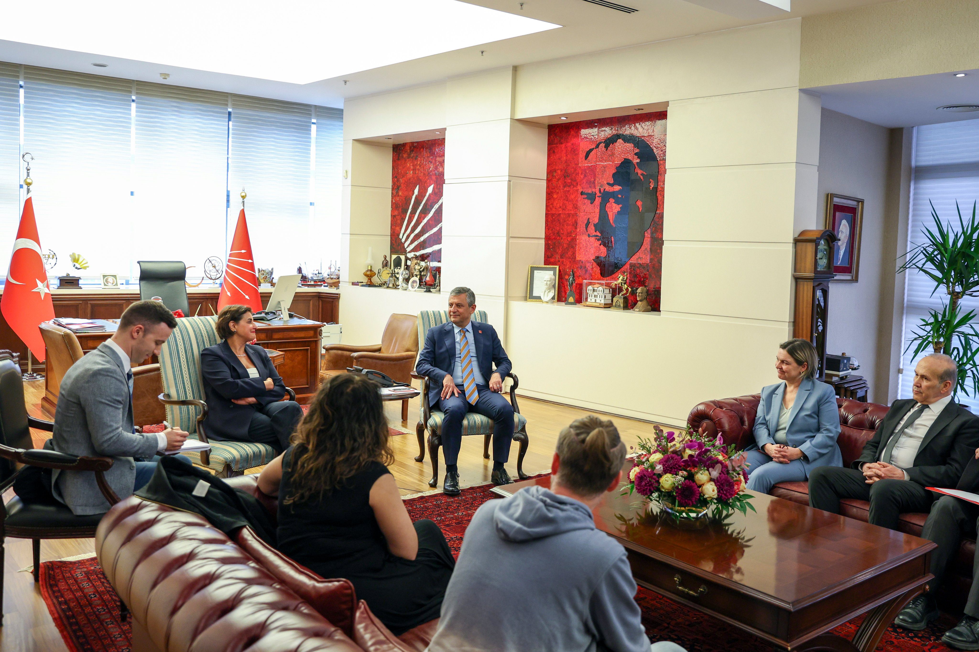 Seven people around a low table with flowers on it, sitting in a large room in comfortable chairs, smiling, with Turkish flags and flags from CHP.